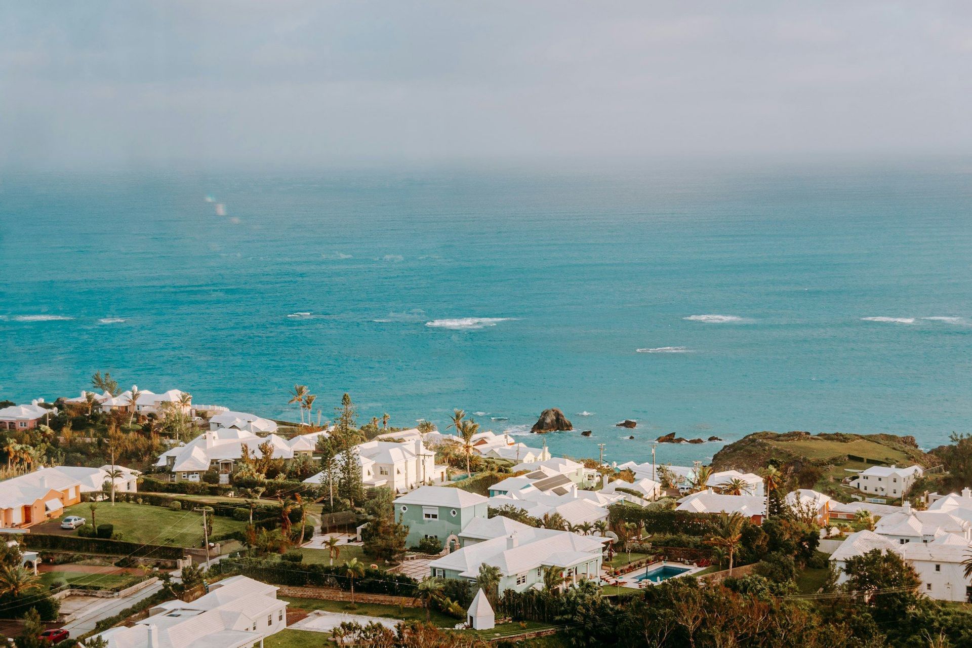 A coastal town with white-roofed houses and lush green vegetation overlooks the turquoise ocean under a hazy sky.