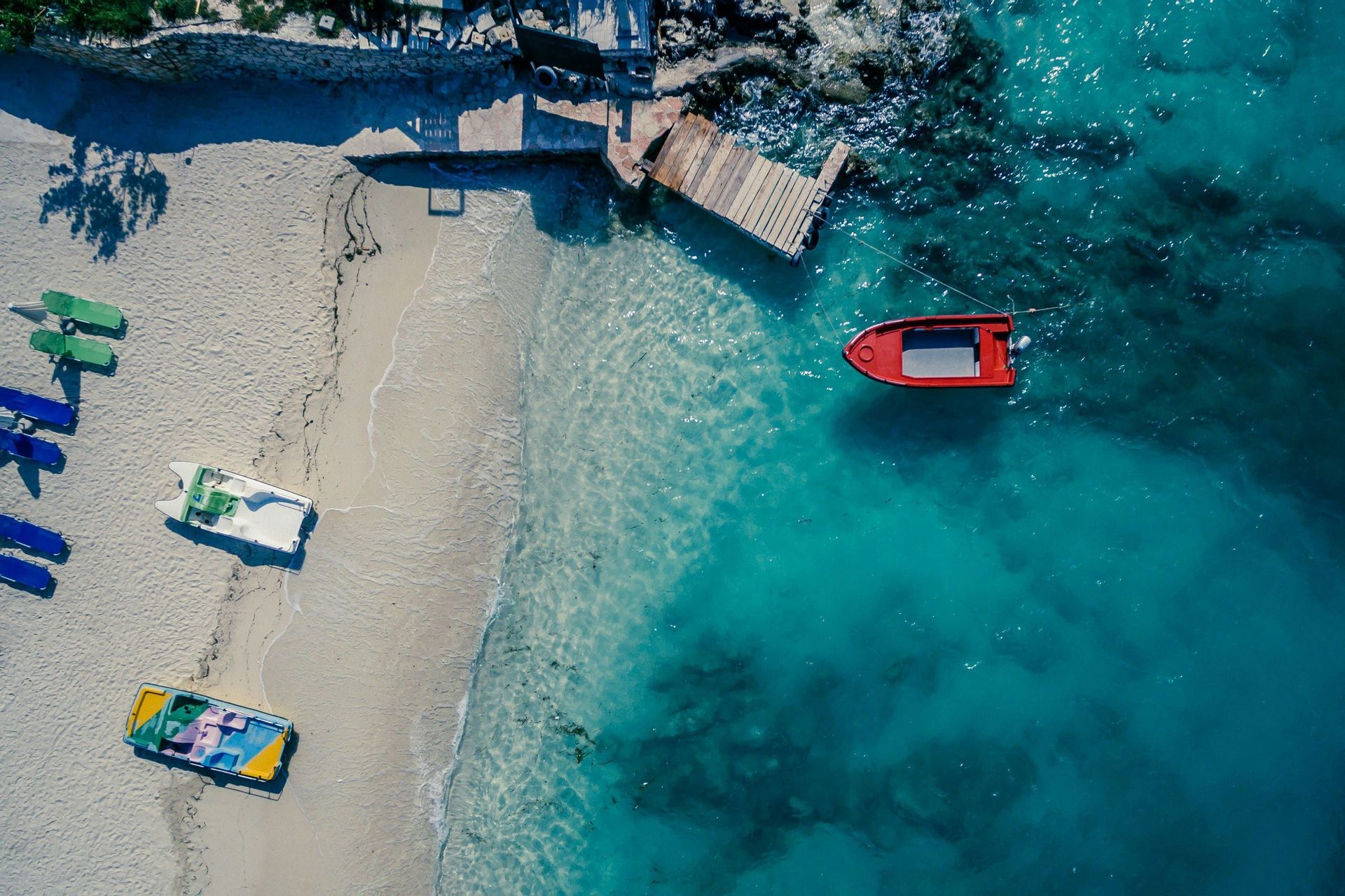 Una vista aérea de una playa de arena con pequeñas embarcaciones en la orilla y un barco rojo flotando en aguas cristalinas de color turquesa junto a un muelle.