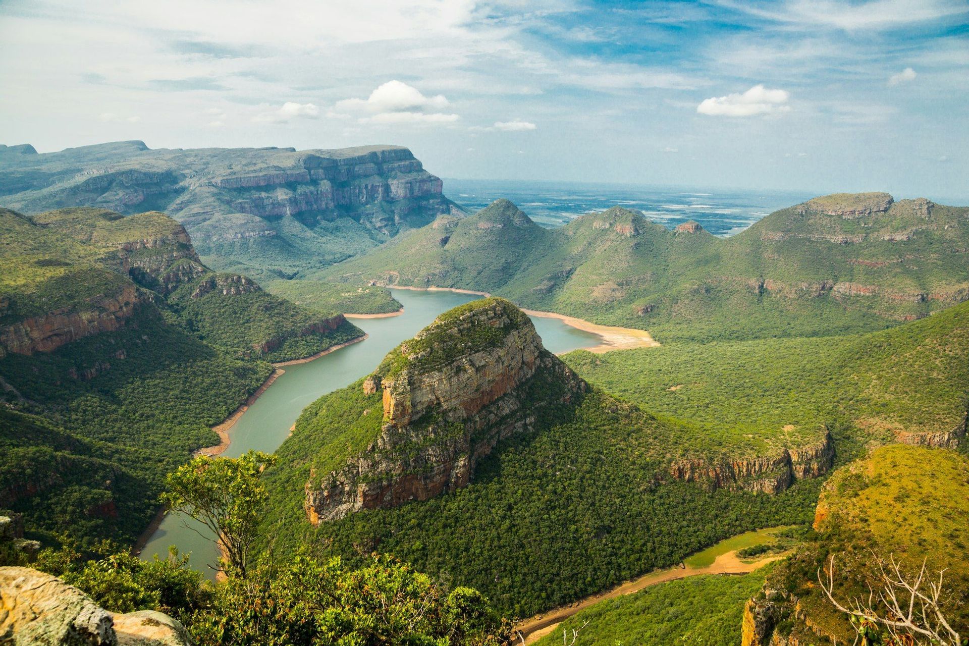 Une vue large et plongeante d'une rivière serpentant à travers un canyon profond rempli de montagnes verdoyantes sous un ciel bleu parsemé de nuages.