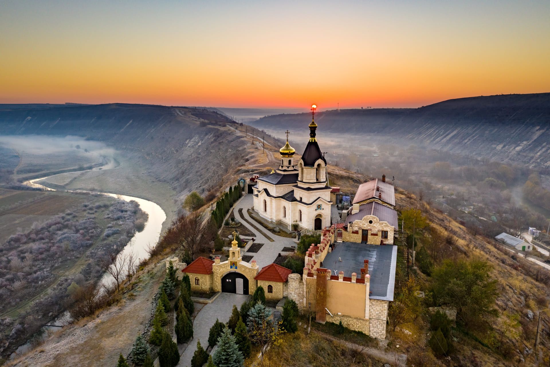 Un monastero ortodosso con cupole dorate sorge su un crinale che domina una valle fluviale nebbiosa all'alba.