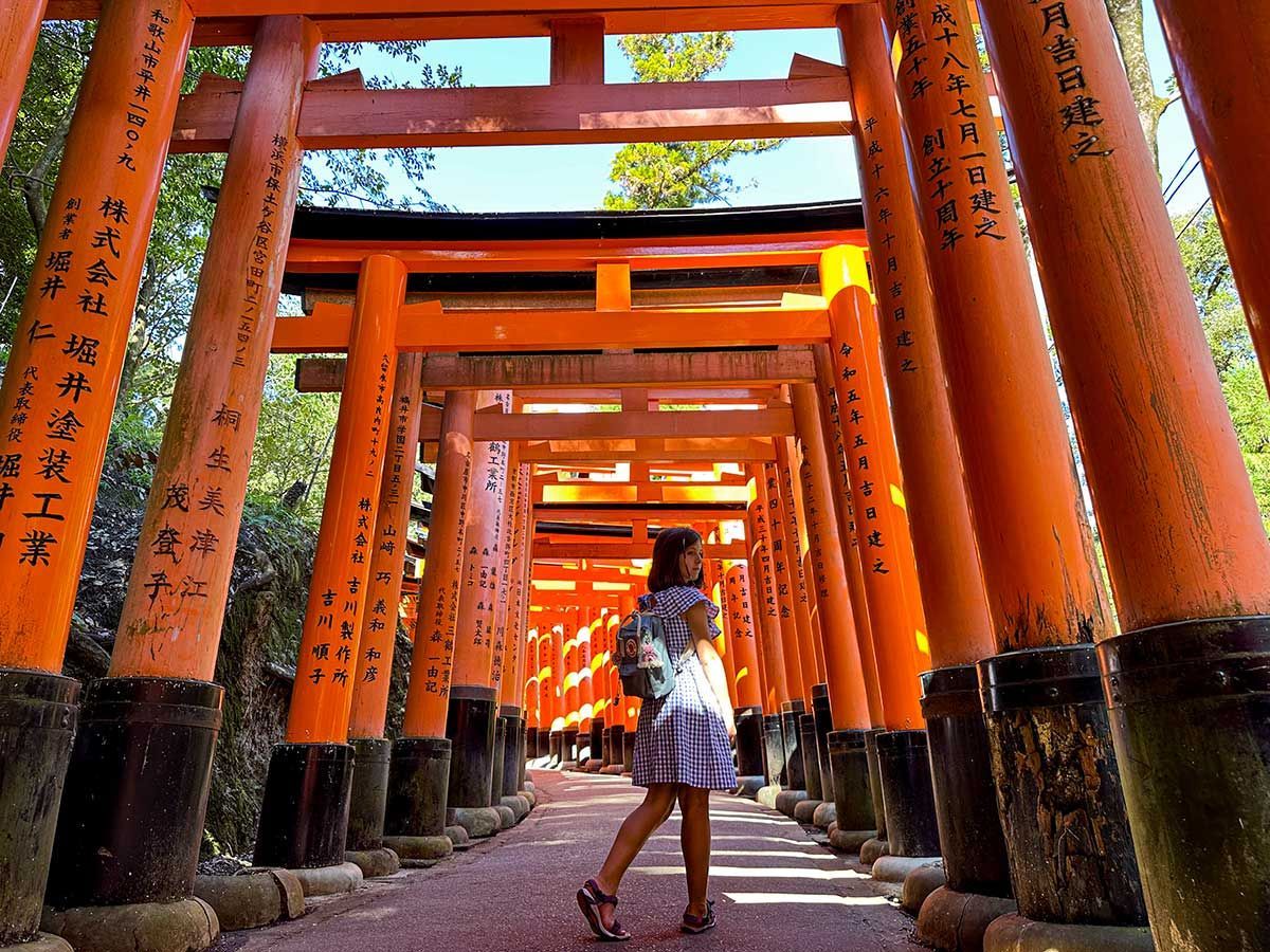 Una donna con uno zaino cammina lungo un sentiero costeggiato da numerosi torii arancioni, voltandosi indietro.