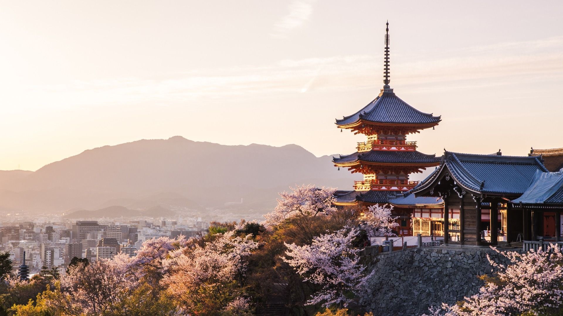 Una pagoda e un tempio giapponese circondati da ciliegi in fiore, affacciati su una città con montagne sullo sfondo al tramonto.