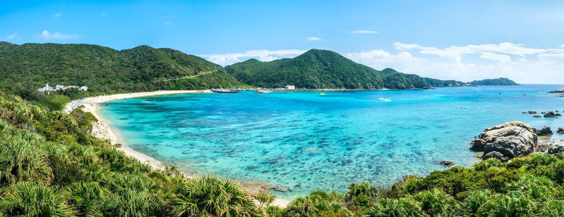 Vista panoramica di una baia tropicale con spiaggia sabbiosa, acqua turchese cristallina e rigogliose colline verdi sotto un cielo azzurro.