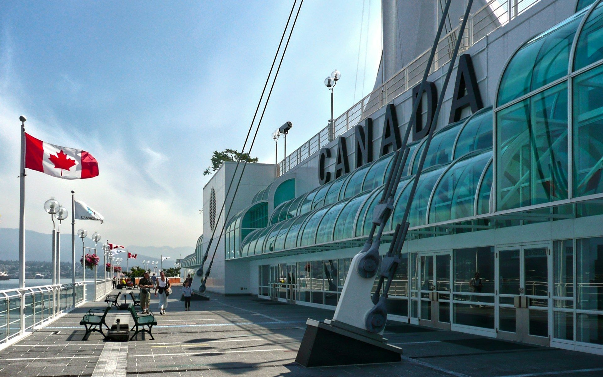 Una bandiera canadese sventola su un molo sul lungomare accanto a un edificio moderno con la scritta 'CANADA' sulla sua facciata sotto un cielo soleggiato.