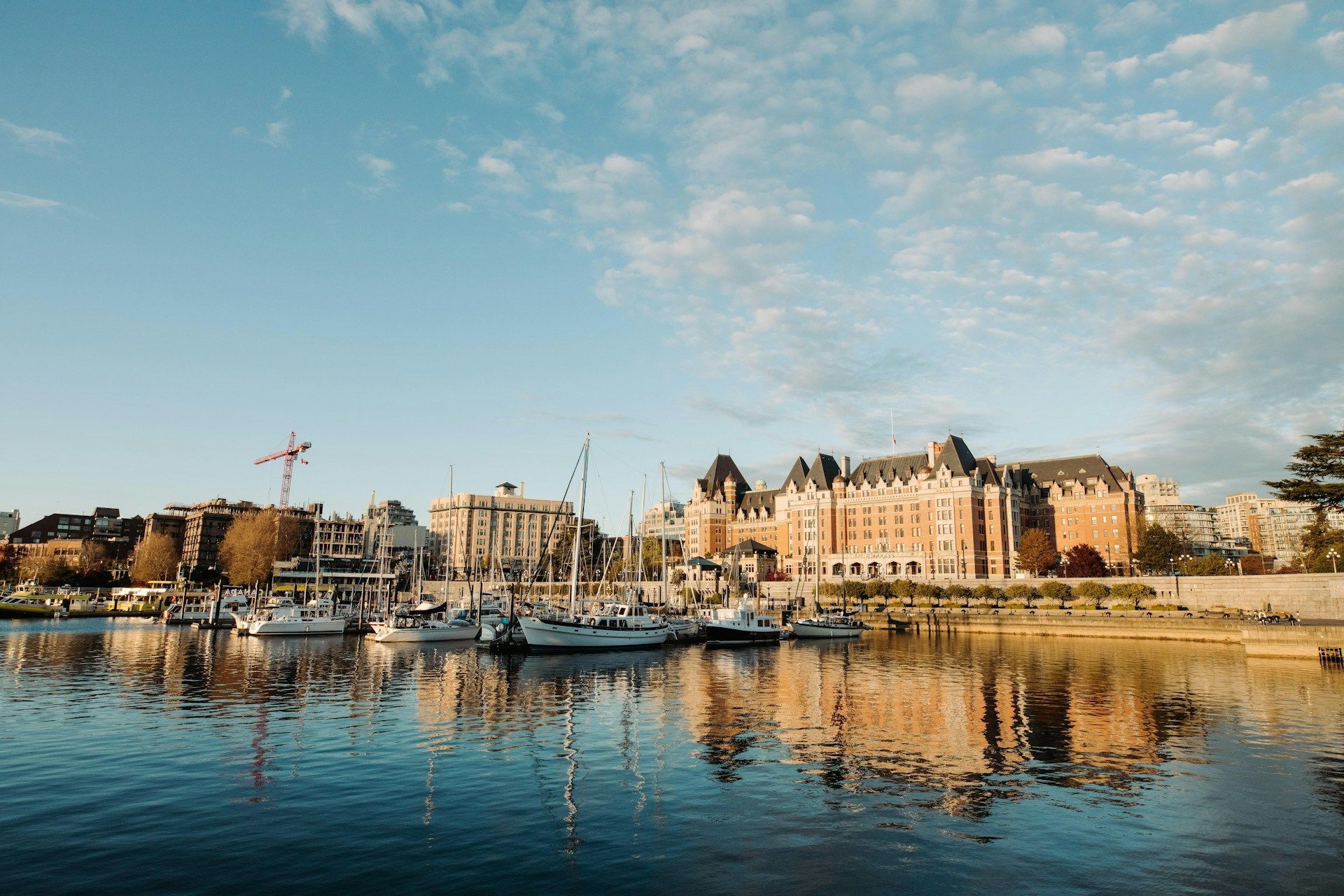 Una vista attraverso un porto calmo mostra barche a vela ormeggiate ed edifici della città riflessi nell'acqua sotto un cielo blu, parzialmente nuvoloso.