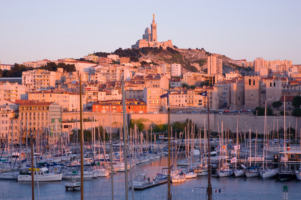 Sailboats docked in a harbor at sunset, with a city and a basilica on a hill in the background.