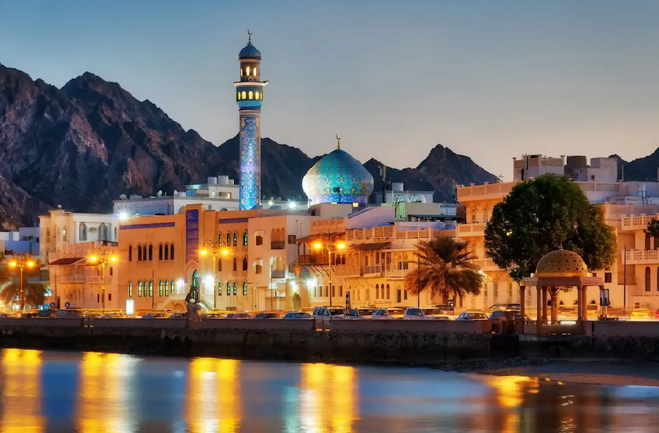 An illuminated mosque with a blue tiled dome and minaret on a city waterfront at dusk, with mountains in the background.