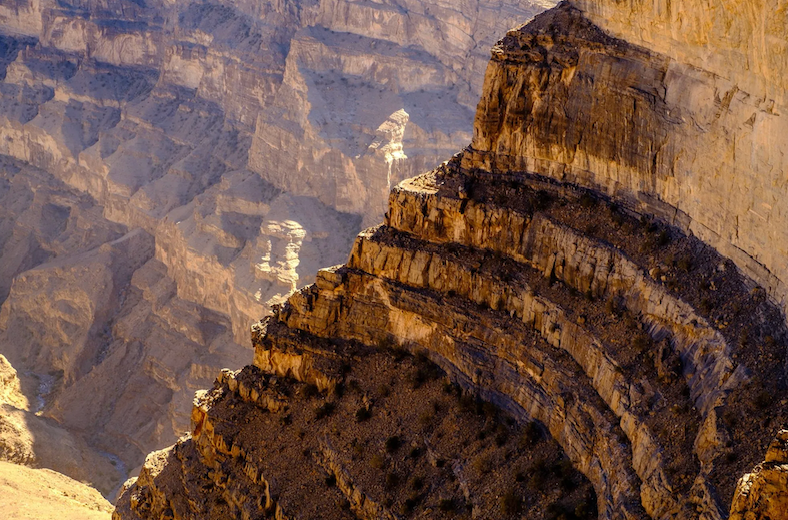 Sunlight illuminates the layered rock strata of a massive canyon wall, with deep shadows in the background.