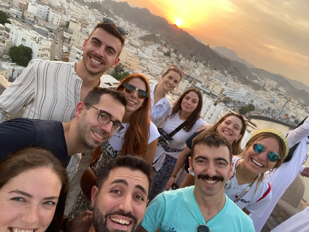 A WeRoad group trip takes a smiling selfie from a high viewpoint, overlooking a coastal city with white buildings at sunset.