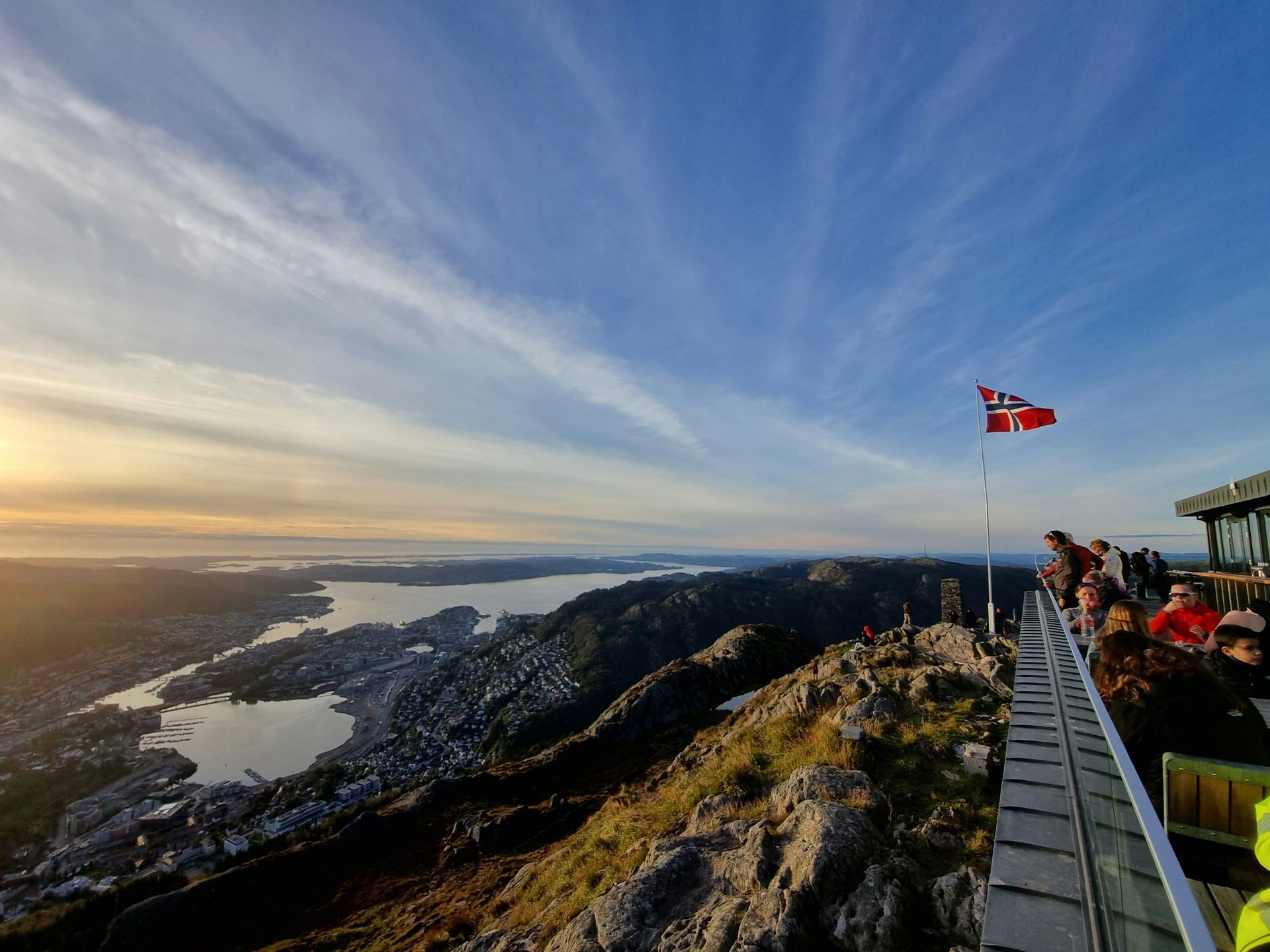 A Norwegian flag flies on a mountain viewpoint where people admire the panoramic view of a coastal city and bay at sunset.