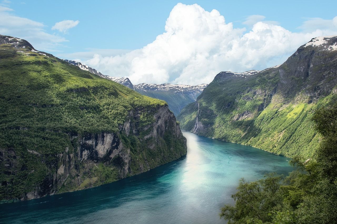 A turquoise fjord winds between steep, forest-covered mountains with snow-capped peaks under a blue sky with white clouds.