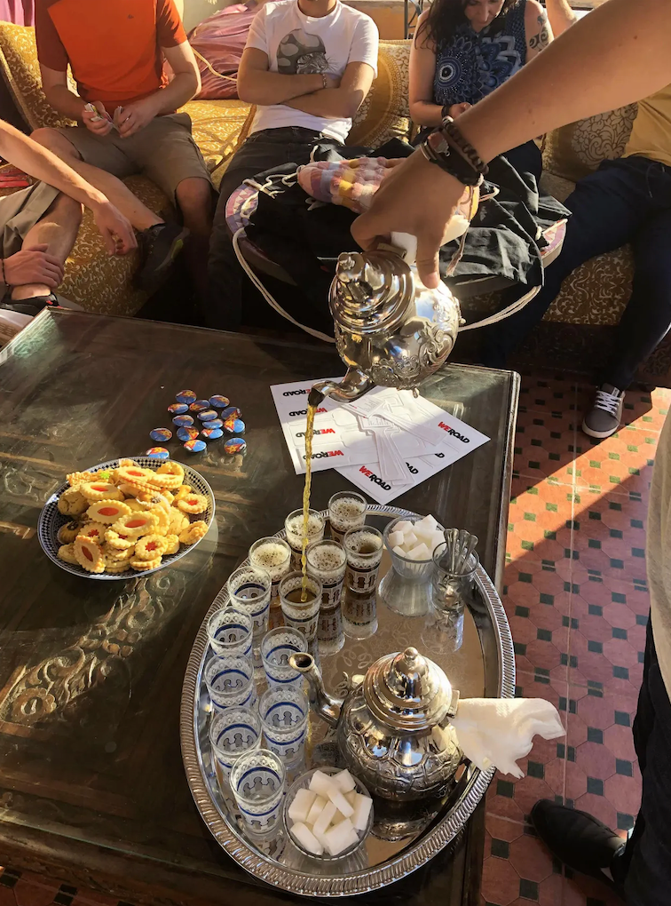 A person pours tea from an ornate silver teapot into small glasses for a WeRoad group trip, with cookies on the table.