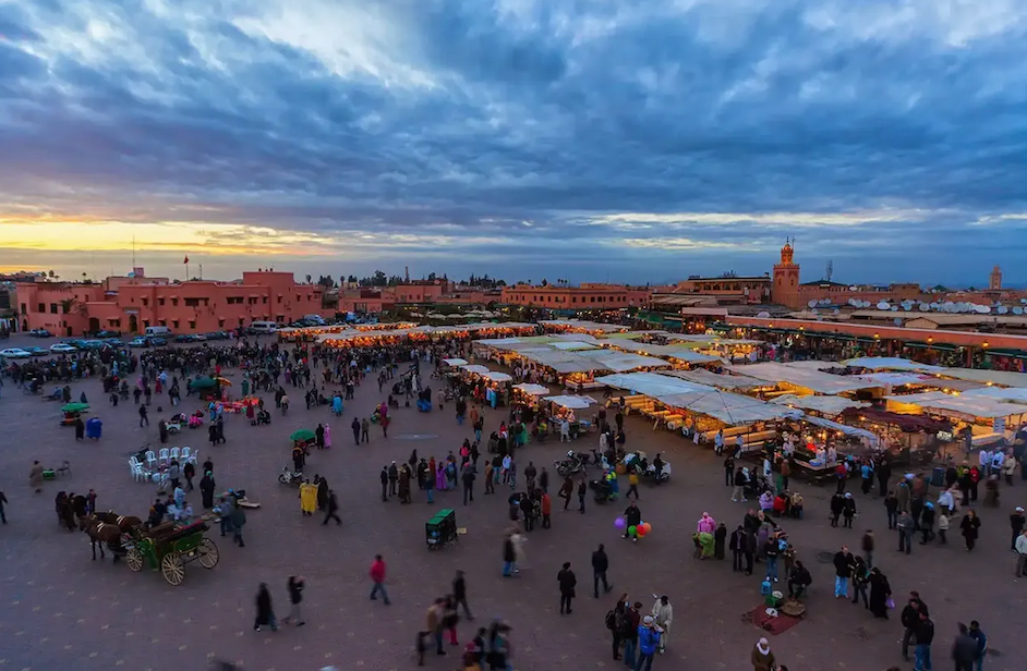 Vue plongeante sur une place de ville animée au crépuscule, remplie de foules et de rangées d'étals de marché illuminés.