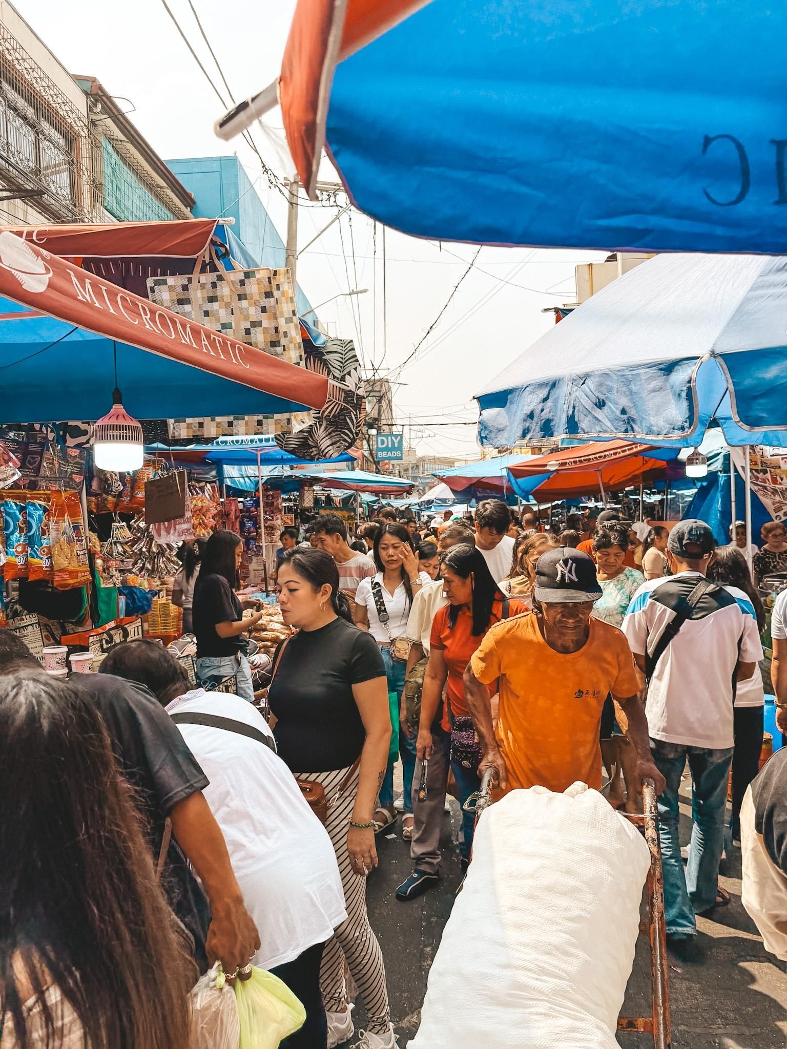 Una folla di persone riempie una strada stretta in un vivace mercato all'aperto, passando tra le bancarelle sotto tende blu e rosse.