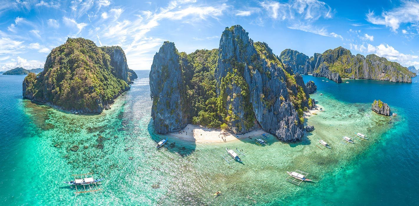 Vista aerea di isole rocciose e ricoperte di vegetazione, con barche a bilanciere su acque limpide e turchesi, vicino a una piccola spiaggia sabbiosa.