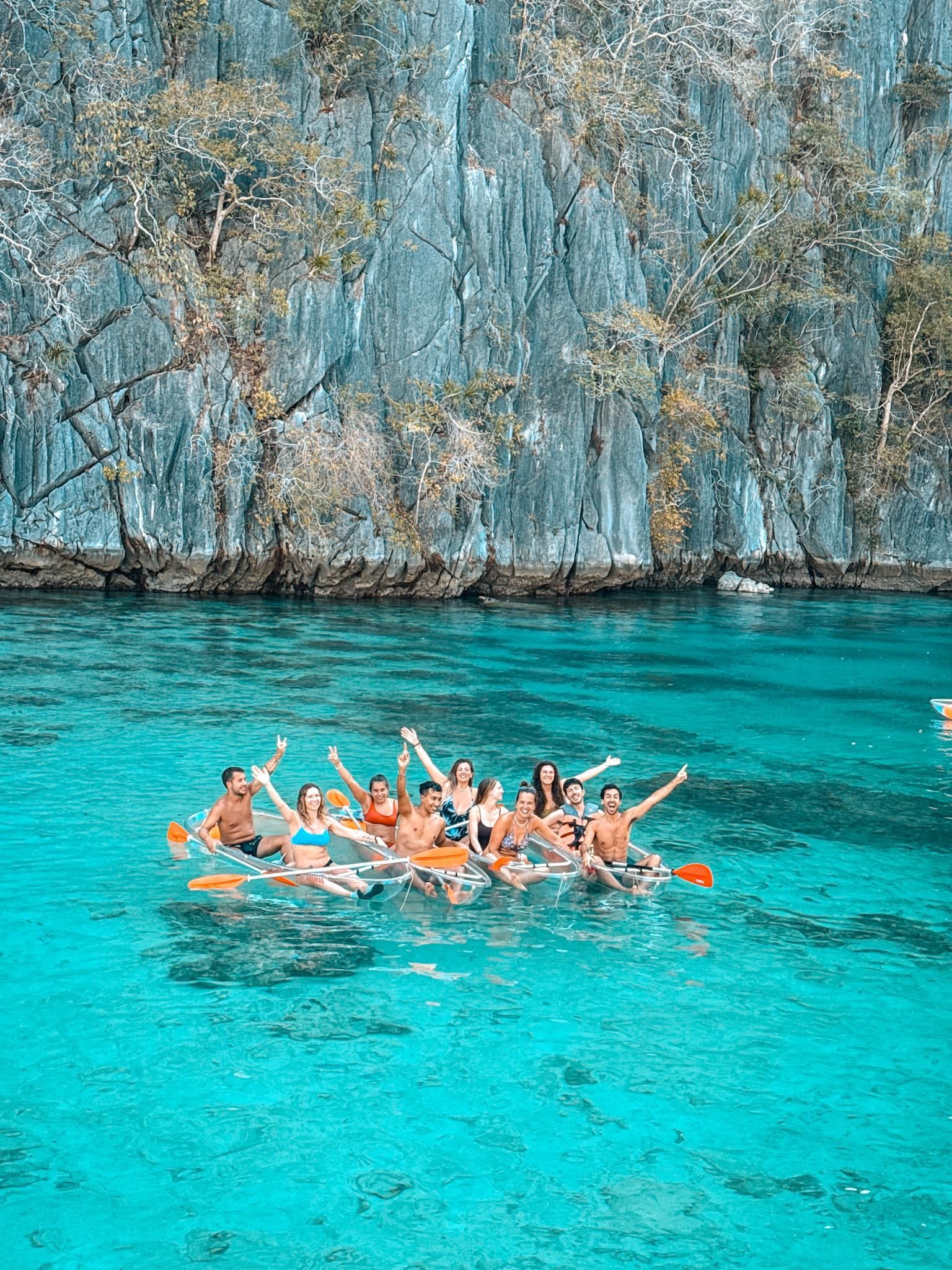 Un gruppo WeRoad sorridente e che saluta da kayak trasparenti su acqua cristallina turchese di fronte a una grande scogliera rocciosa.