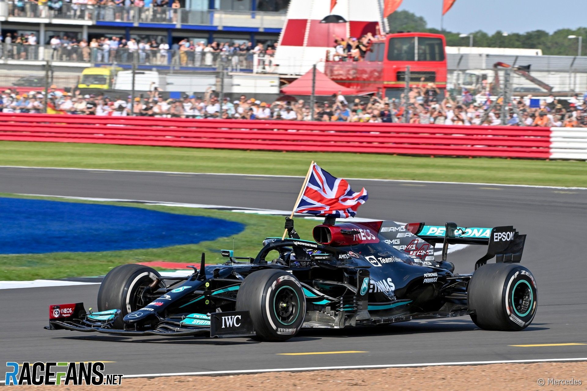 A black Formula 1 car with a British flag drives on a racetrack as spectators watch from the grandstands in the background.