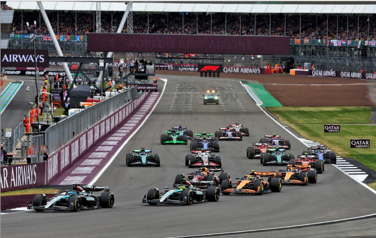 A pack of Formula 1 cars race down a track with grandstands full of spectators in the background.