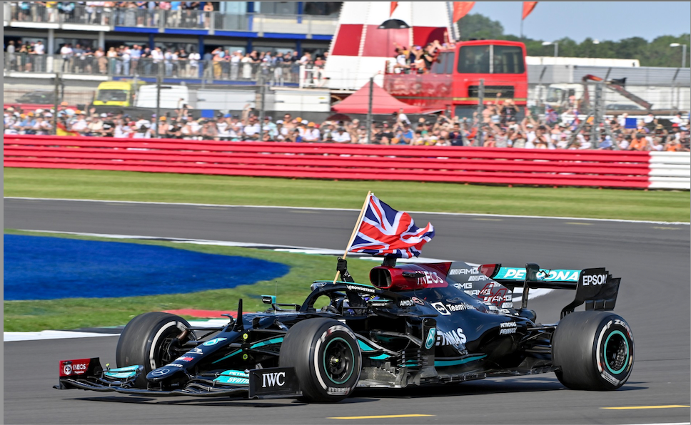 A black Formula 1 car with a British flag attached drives along a racetrack in front of a crowd of spectators in the stands.