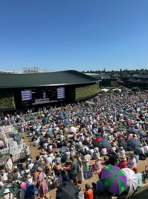 A large crowd of people sitting on a grassy hill watching a tennis match on a big screen at an outdoor venue on a sunny day.