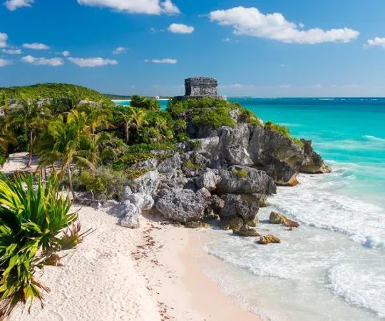 An ancient stone ruin sits atop a rocky cliff covered in palm trees, overlooking a white sand beach and turquoise sea.