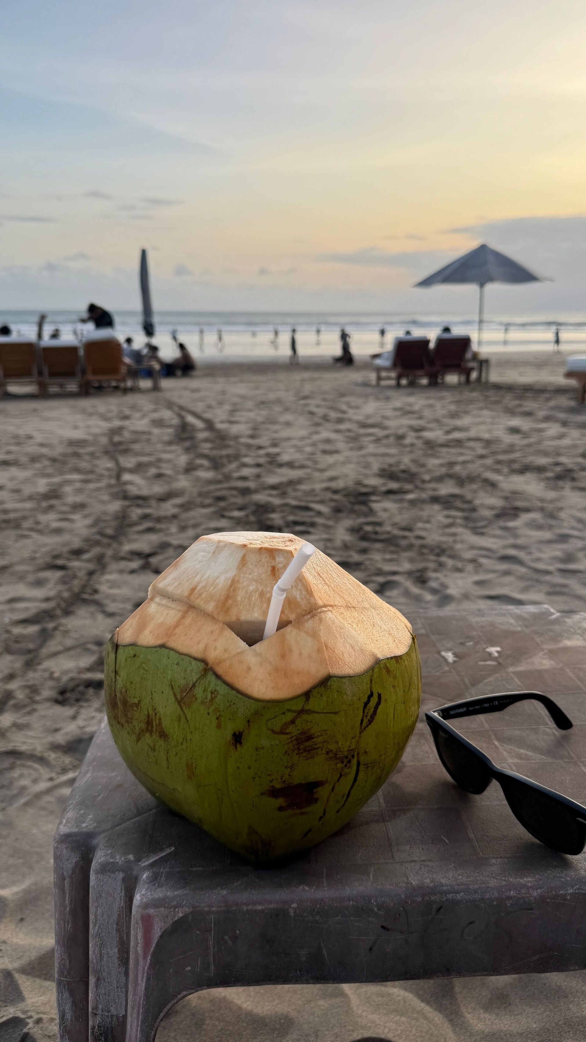 Un cocco verde con una cannuccia su un tavolo accanto a occhiali da sole su una spiaggia sabbiosa al tramonto.