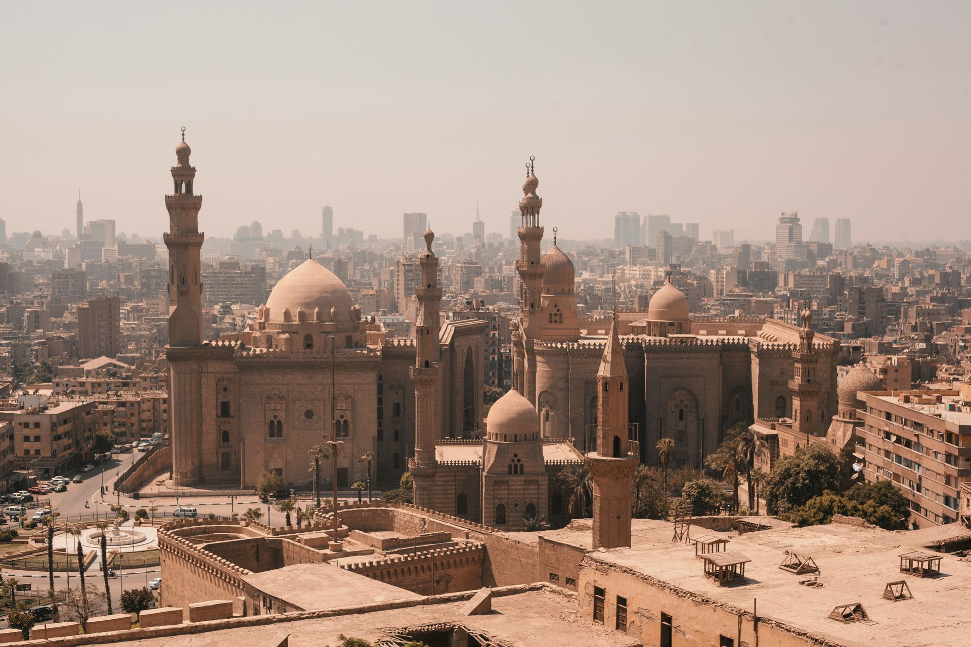 An aerial view of a large, sand-colored mosque with multiple domes and minarets against a hazy cityscape with modern skyscrapers in the background.