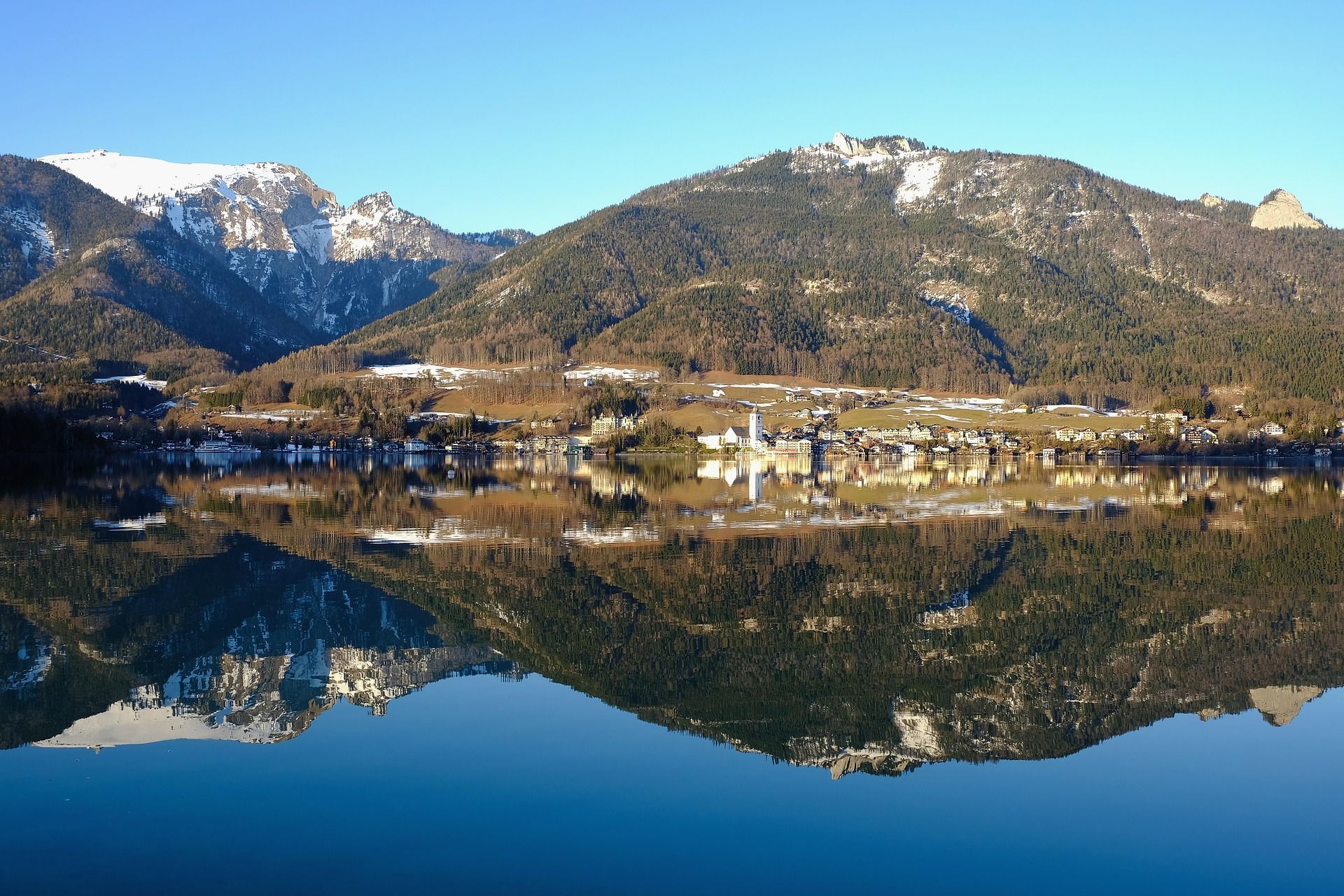 Un borgo lacustre e montagne innevate che si riflettono perfettamente nelle acque calme sotto un cielo azzurro.