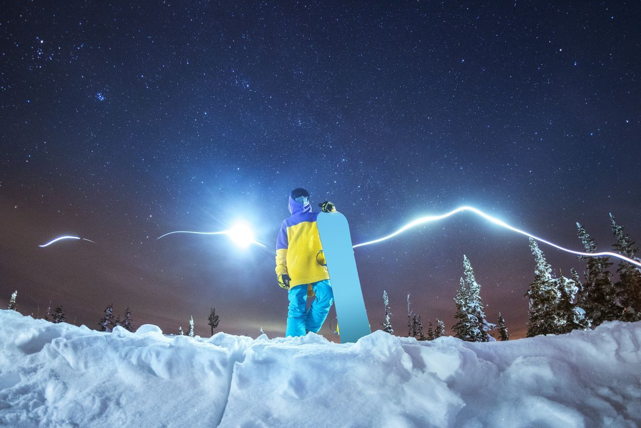 Uno snowboarder con giacca gialla tiene uno snowboard, in piedi su un paesaggio innevato sotto un cielo notturno stellato con scie luminose.