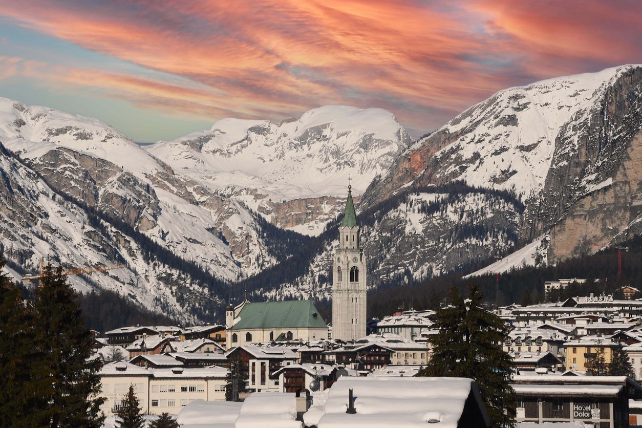 Un villaggio innevato con un'imponente torre di chiesa alla base di grandi montagne coperte di neve sotto un cielo drammatico rosa e arancione.