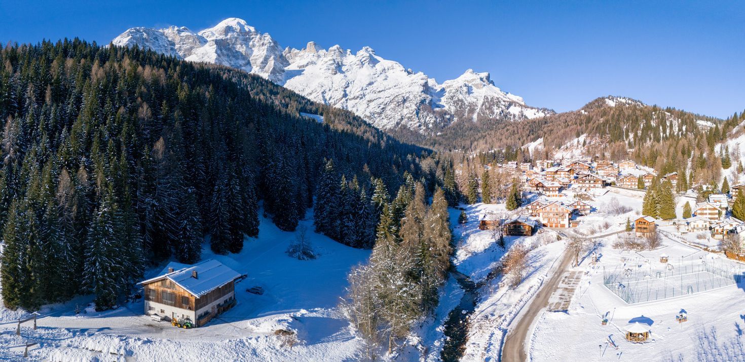 Una veduta aerea di un villaggio di montagna innevato immerso in una pineta, con grandi cime innevate sullo sfondo sotto un cielo azzurro.