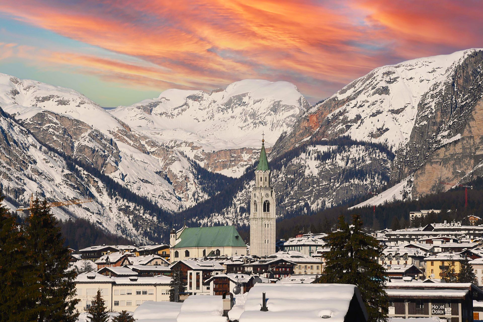 A snow-covered village with a tall church bell tower nestled against massive, snowy mountains under an orange and pink sky.
