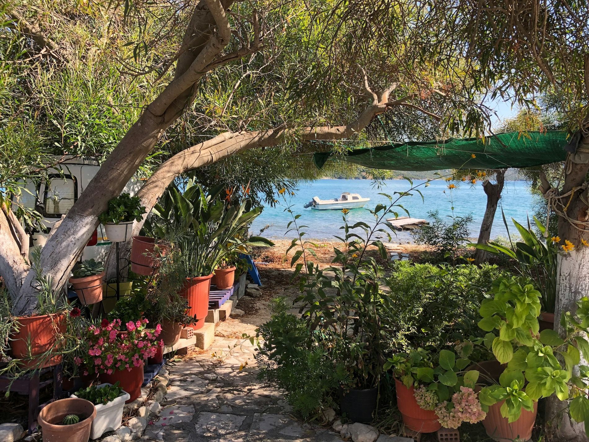 Un sentiero in pietra attraverso un lussureggiante giardino ricco di piante in vaso e alberi conduce a una baia calma e blu con una piccola barca a motore.