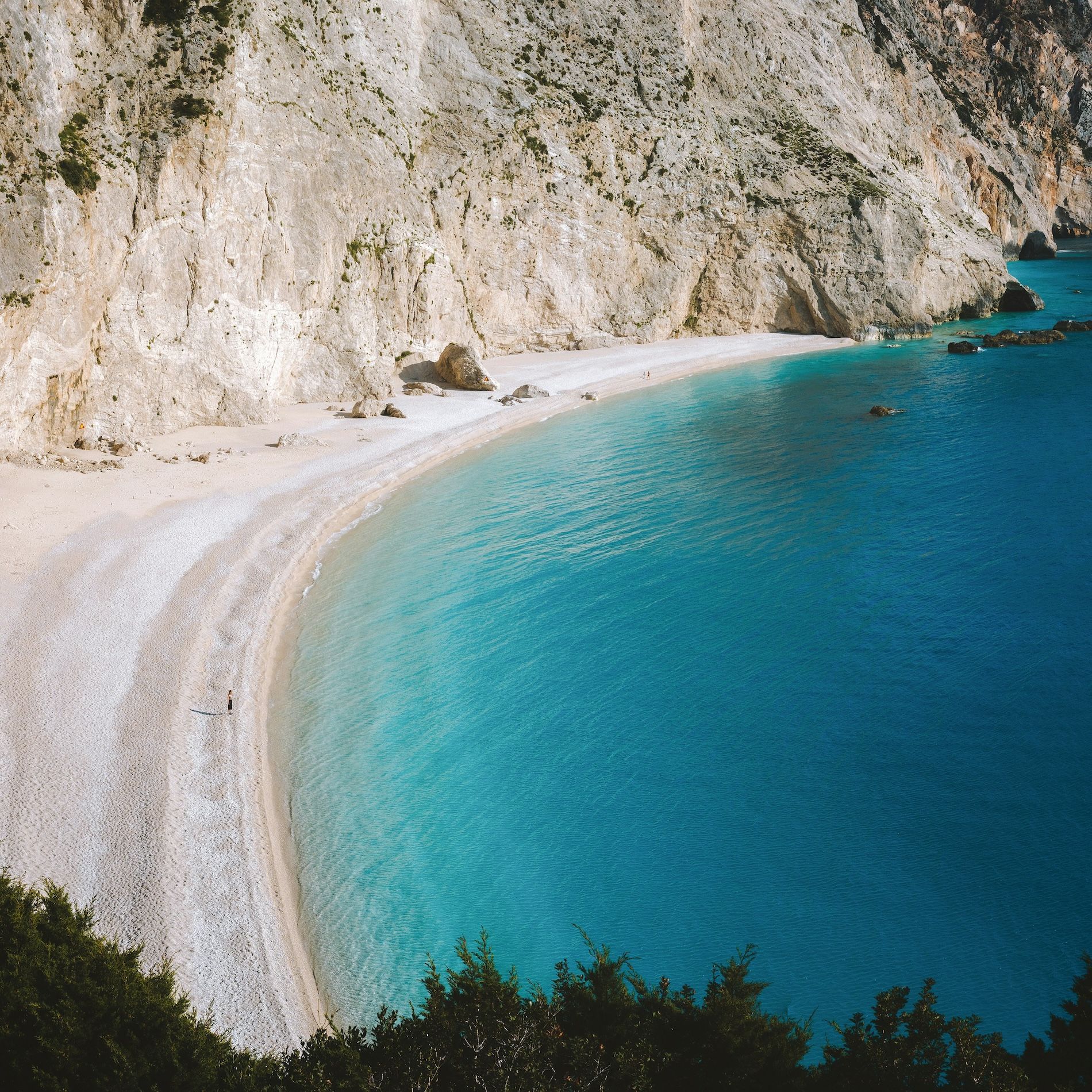 Una vista aerea di una spiaggia bianca e appartata, con acque turchesi e vibranti, incastonata ai piedi di un'ampia scogliera rocciosa.
