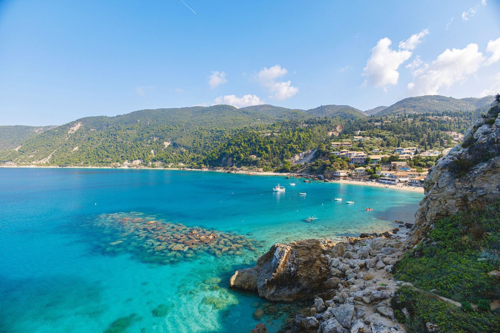 Una vista dall'alto di una baia costiera con acqua cristallina color turchese, una spiaggia sabbiosa e un piccolo villaggio immerso nelle verdi colline.