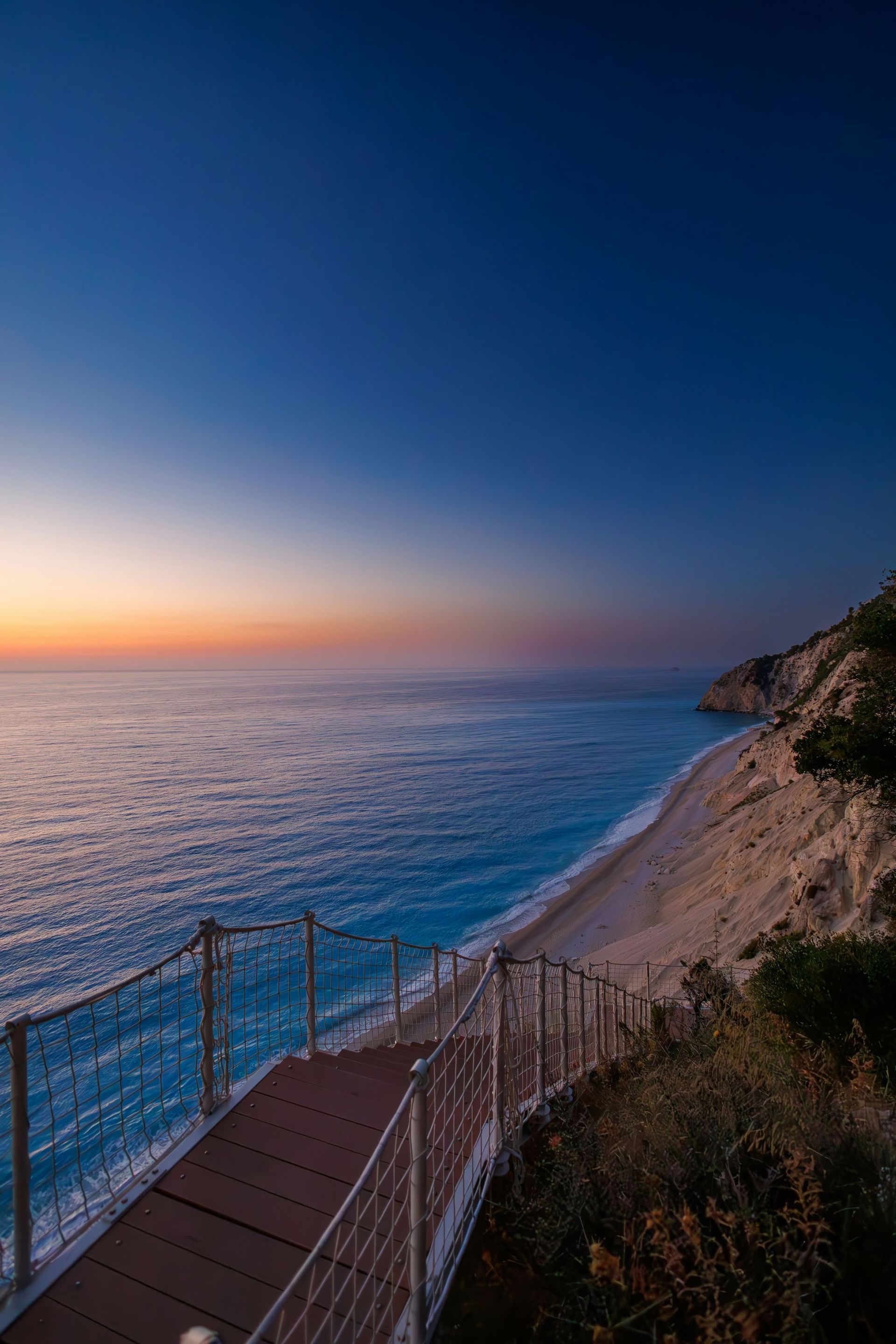 Una scala di legno con ringhiera in corda e rete scende da una scogliera verso una spiaggia sabbiosa e un mare turchese al tramonto.