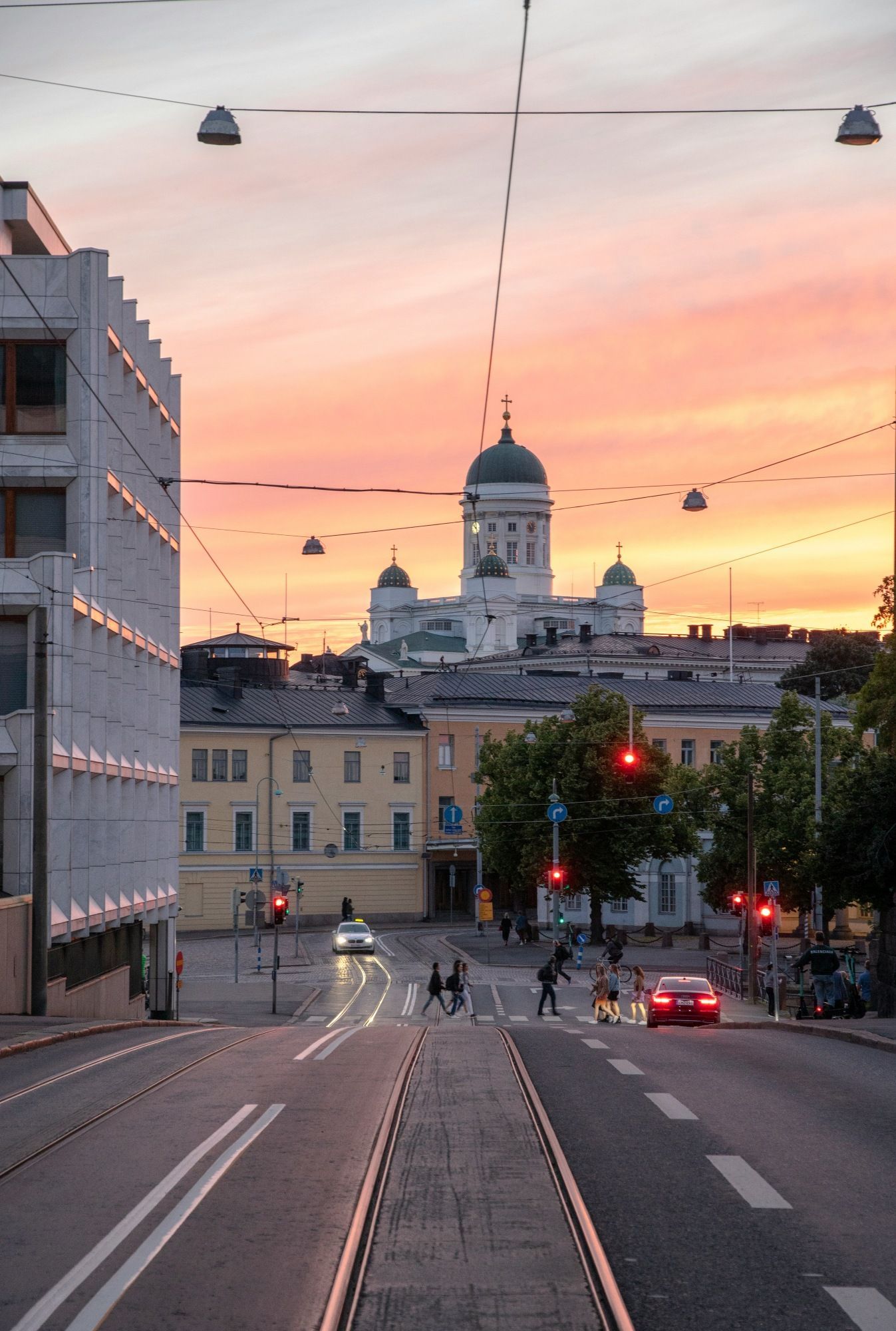 Blick auf eine Stadtstraße mit Straßenbahnschienen und einer großen weißen Kathedrale mit grünen Kuppeln, die sich vor einem Sonnenuntergangshimmel abzeichnet.