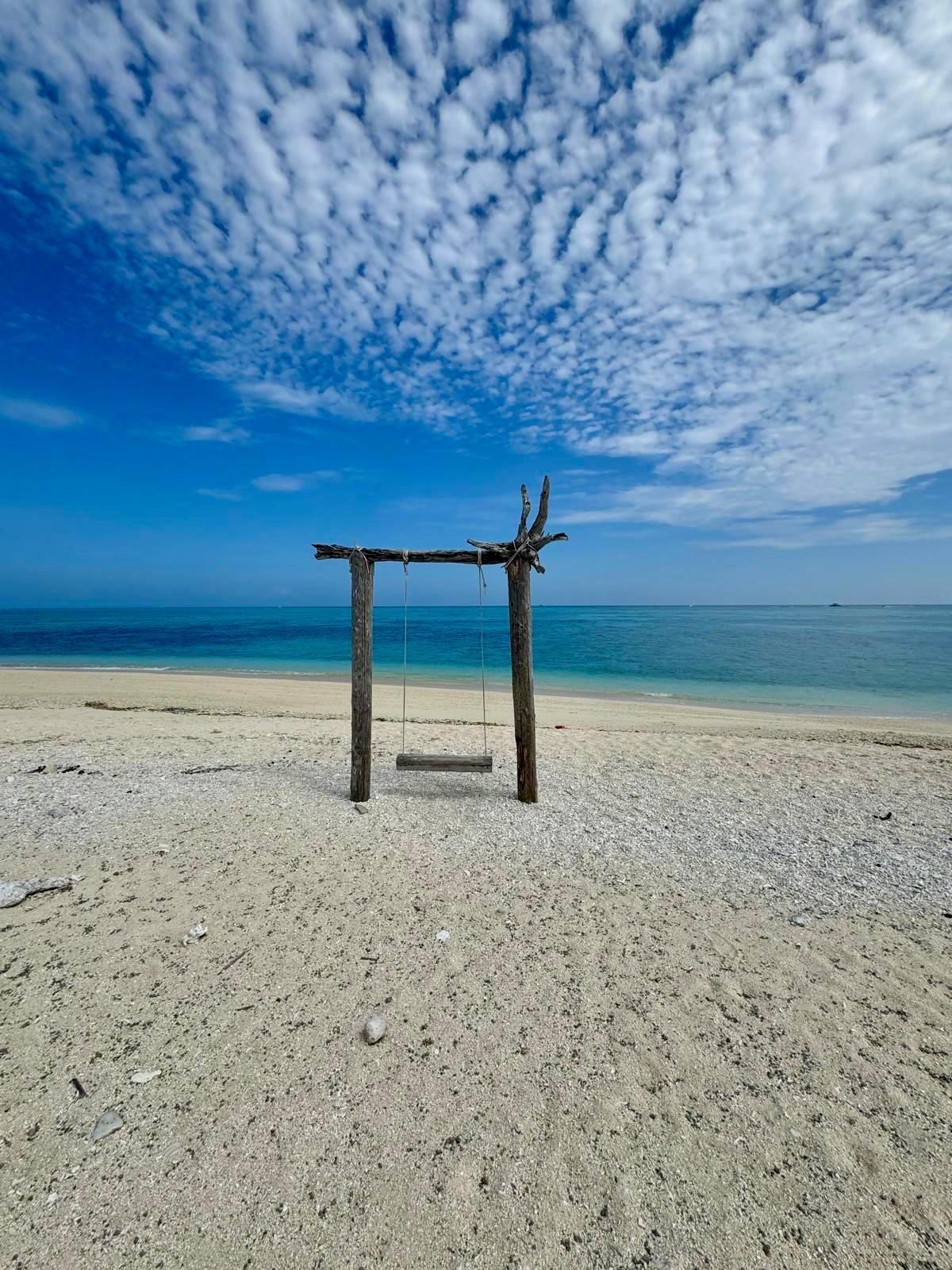 Un'altalena rustica in legno, vuota, su una spiaggia sabbiosa, di fronte all'oceano calmo e blu sotto un cielo parzialmente nuvoloso.