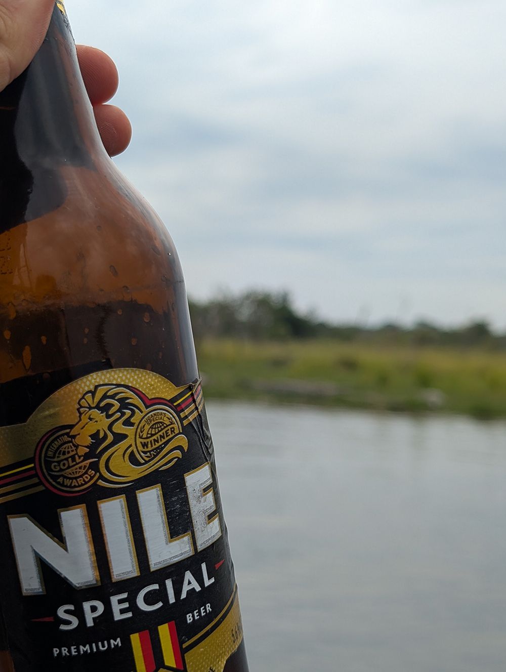 Primer plano de una mano sosteniendo una botella de cerveza Nile Special con un río y un paisaje verde de fondo.