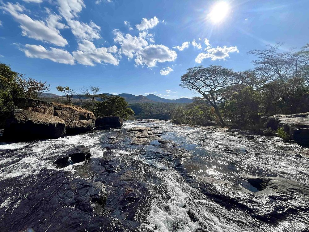 Un fiume ampio e roccioso scorre rapidamente sotto un sole splendente e un cielo azzurro nuvoloso, con colline boscose in lontananza.