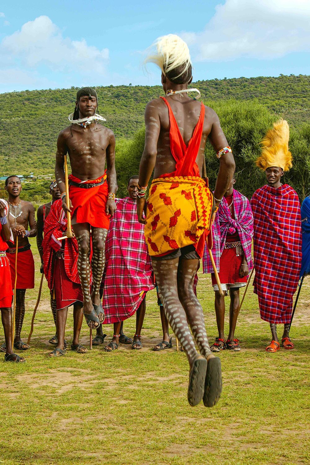 Due uomini Maasai in abiti tradizionali saltano in alto durante una danza cerimoniale, mentre altri uomini osservano su una pianura erbosa.
