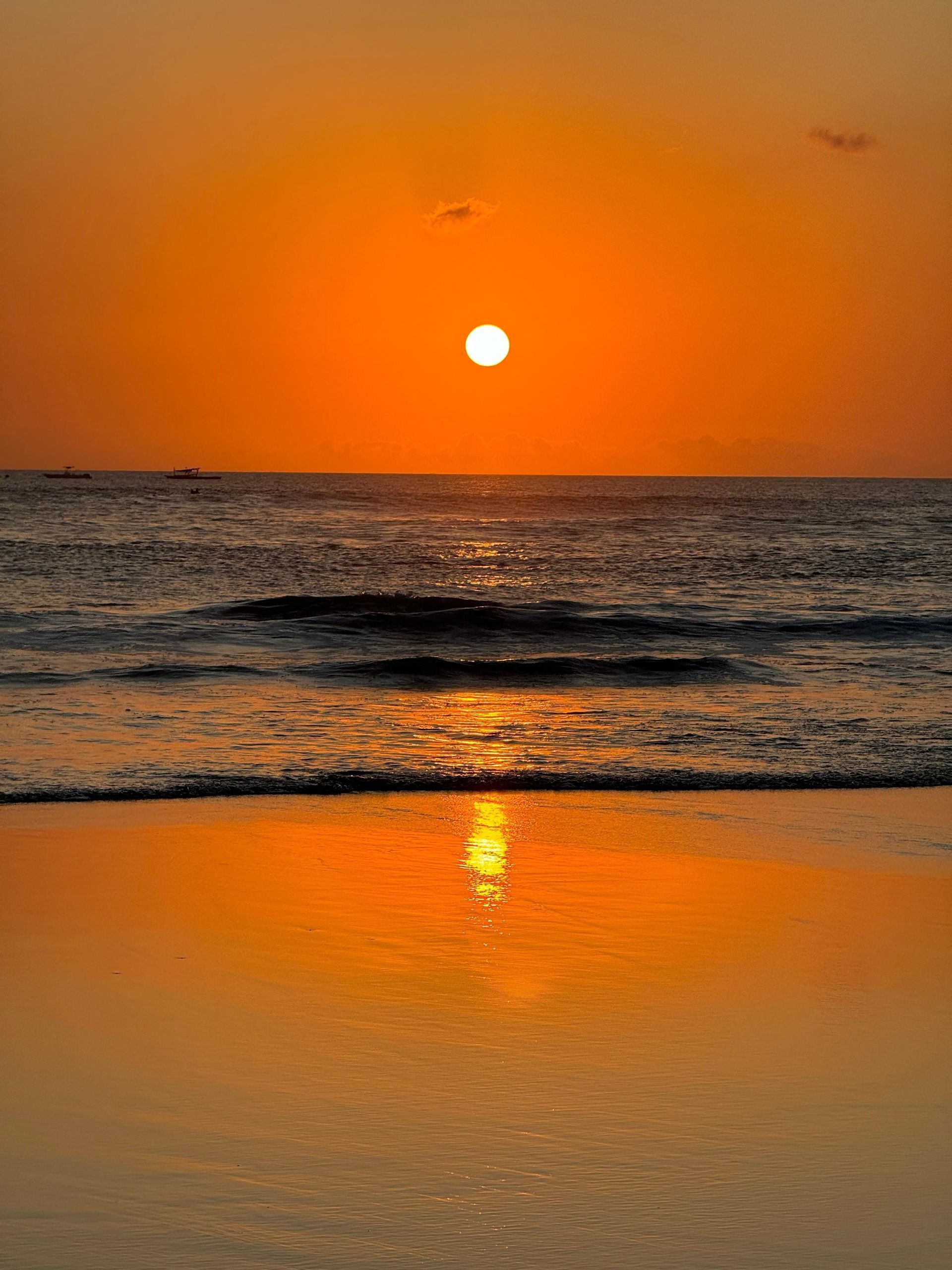 Il sole al tramonto brilla in un cielo arancione, la sua luce si riflette sull'oceano e sulla sabbia bagnata di una spiaggia.
