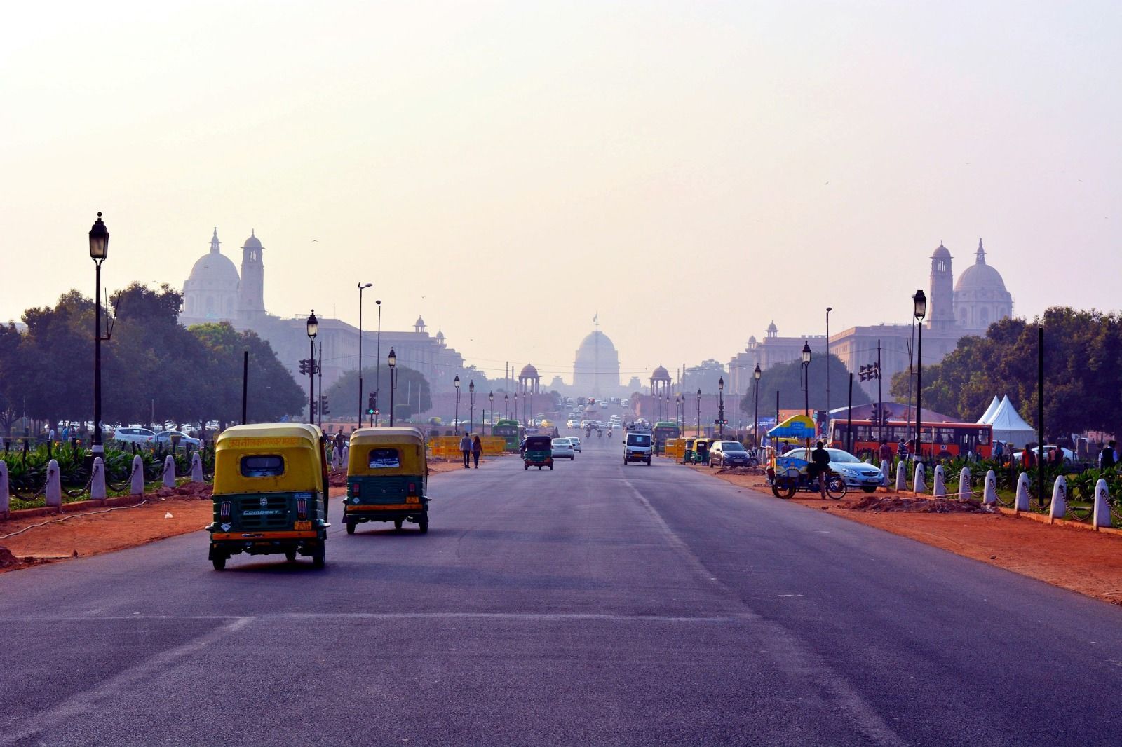 Two yellow and green auto-rickshaws driving on a wide road leading towards large domed buildings under a hazy sky.