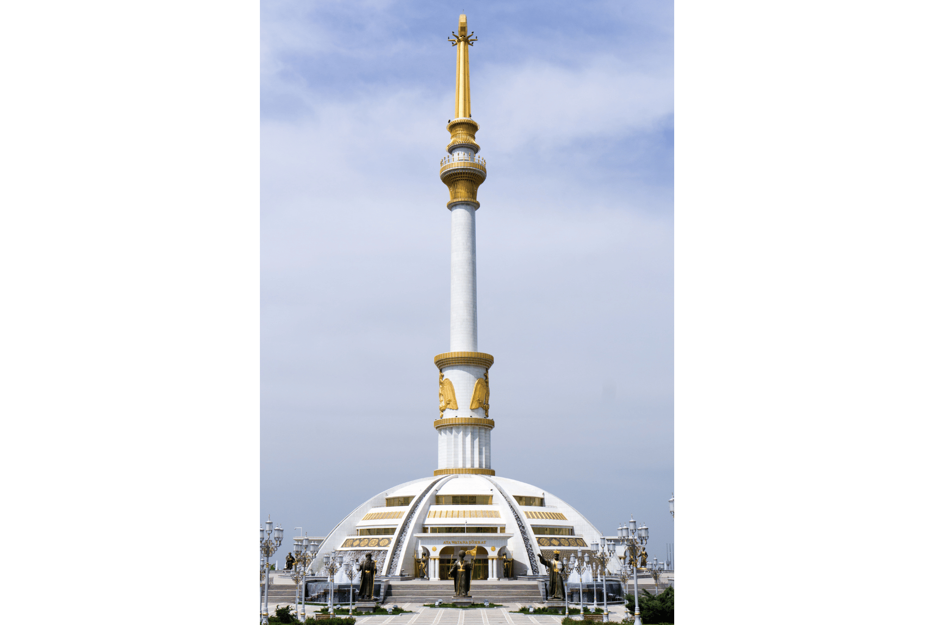 Un alto monumento bianco, con guglia e dettagli dorati, si erge su una base a cupola in un'ampia piazza sotto un cielo nuvoloso.