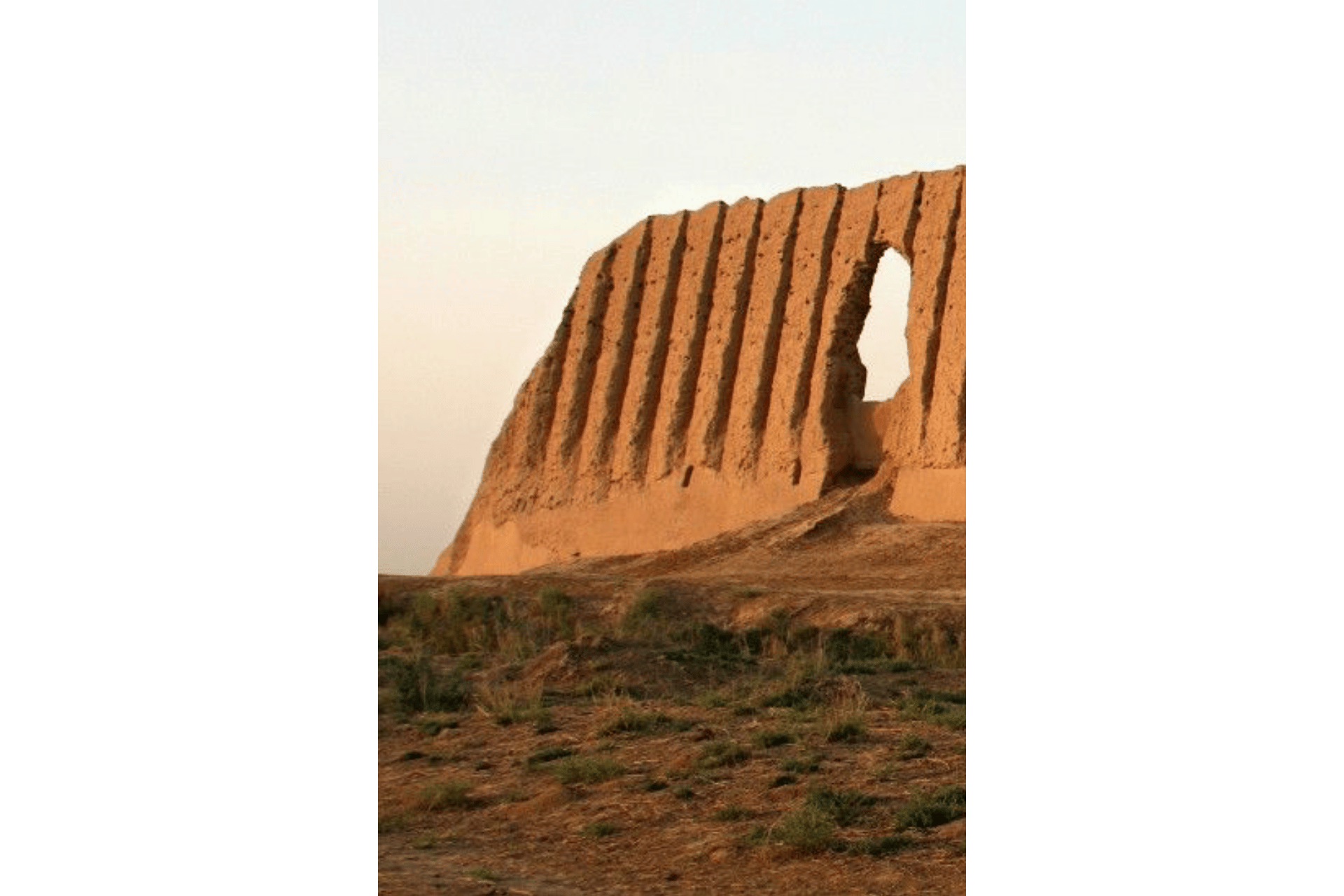 Una rovina di muro in mattoni di fango ondulato con una grande apertura si erge su una collina secca ed erbosa contro un cielo pallido e velato.