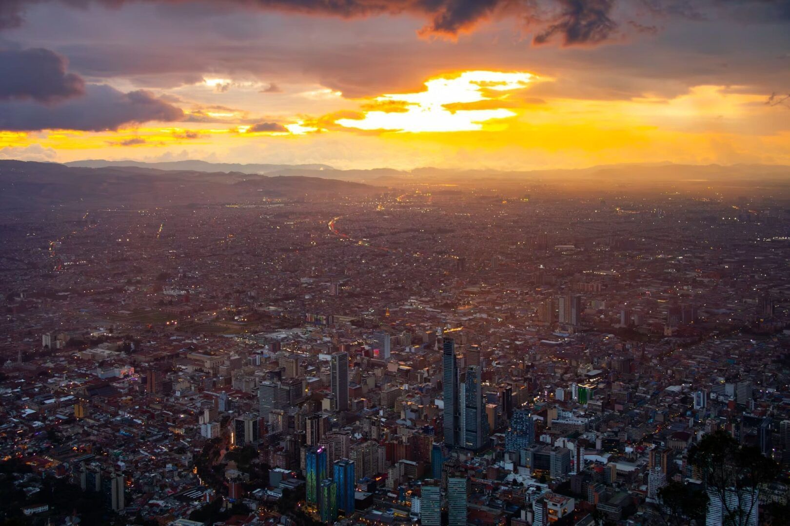 Una vista aerea di un'estesa città con edifici illuminati al tramonto, sotto un cielo drammatico con un brillante tramonto dorato.