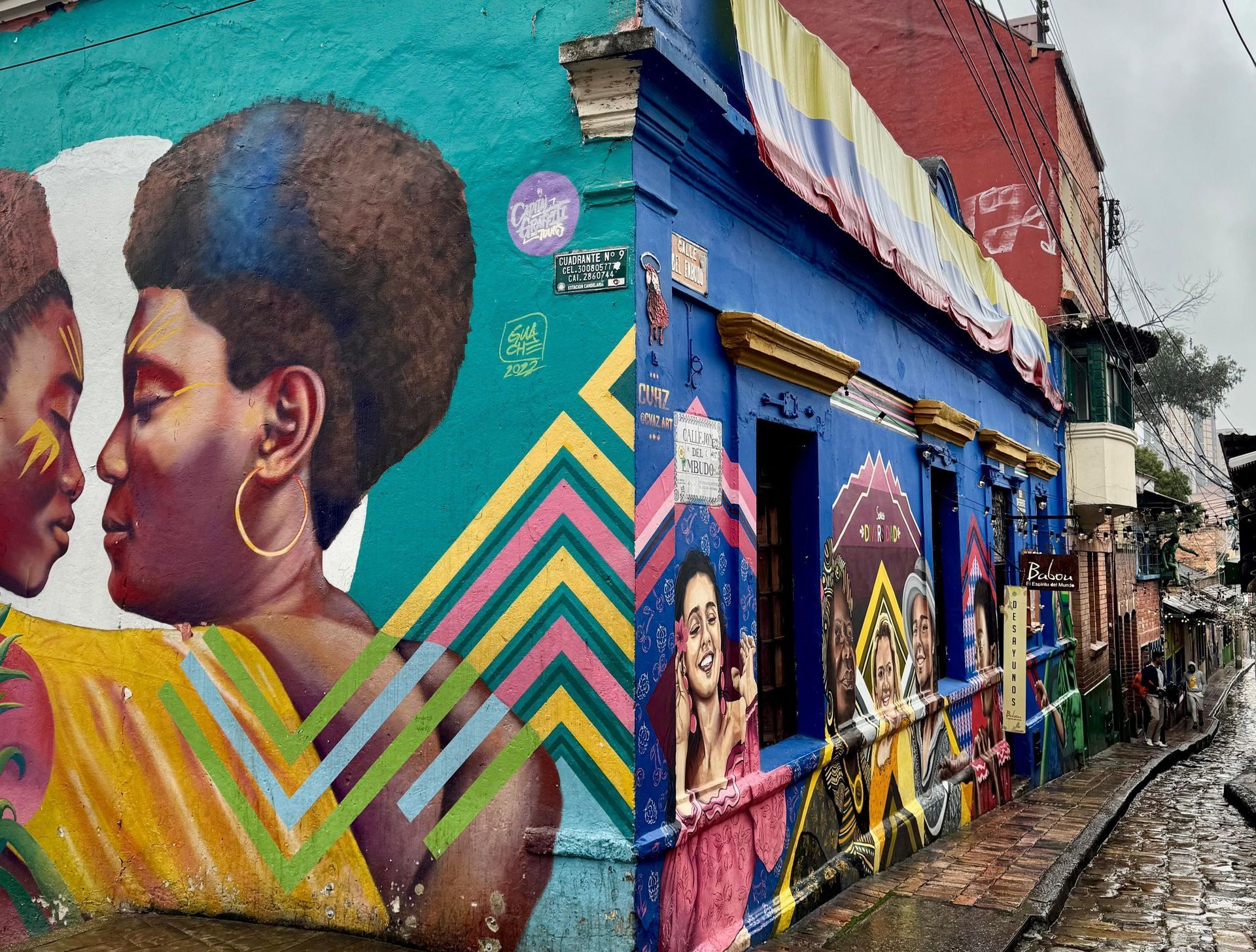 Un edificio d'angolo con murales colorati e una bandiera colombiana appesa, su una stretta strada acciottolata e bagnata.