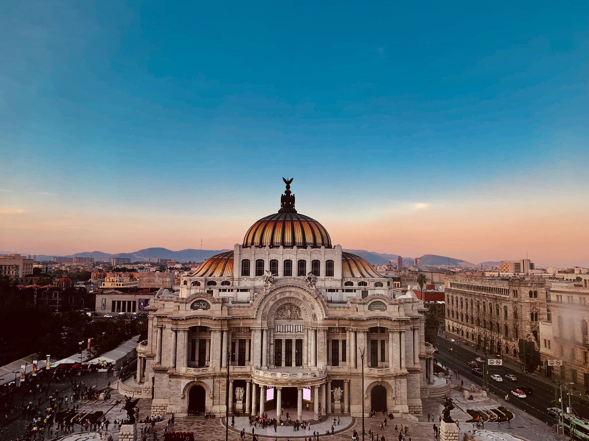 Un edificio decorato con cupola si erge in una vivace piazza cittadina al tramonto, con uno skyline e montagne visibili sullo sfondo.
