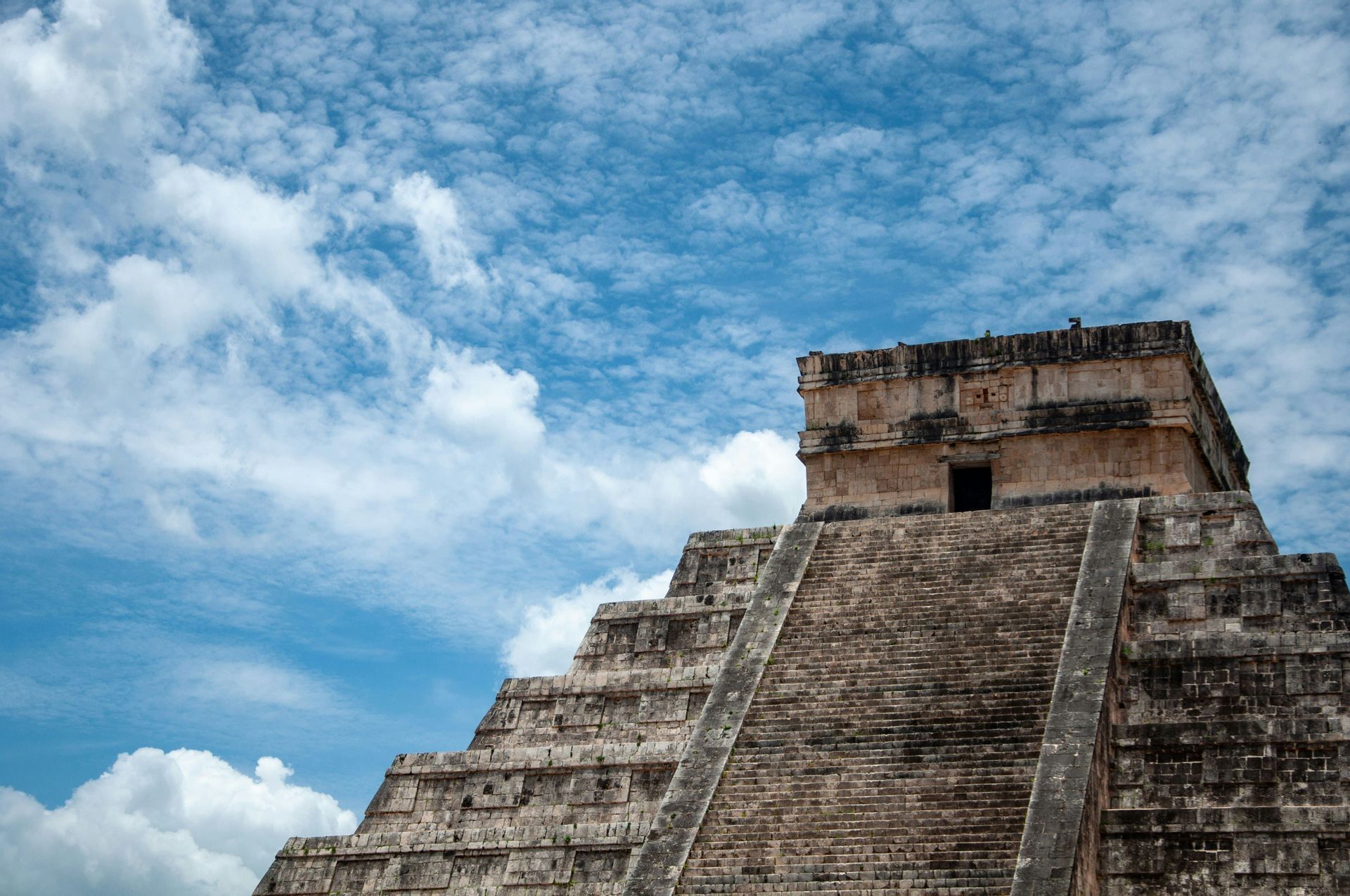 Una vista dal basso di un'antica piramide a gradoni in pietra che si eleva verso un cielo blu con nuvole bianche sparse.