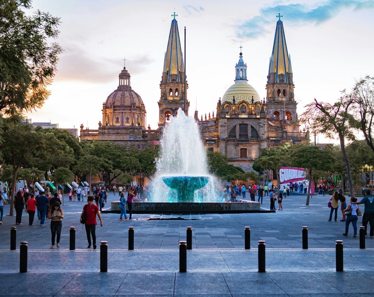 Una grande fontana spruzza acqua in una vivace piazza cittadina di fronte a una cattedrale ornata con guglie gemelle al tramonto.